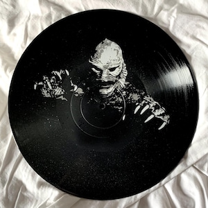 May include: A black vinyl record with a white silhouette of the Creature from the Black Lagoon. The creature's face is visible, with sharp teeth and a menacing expression.