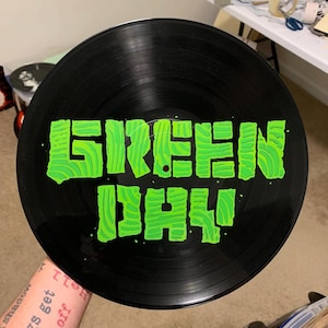 May include: A black vinyl record with the words "GREEN DAY" painted in bright green. The lettering has a textured, layered appearance. The record is held up, with the artist's name prominently displayed.