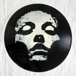 May include: A black vinyl record with a white, stylized face design. The face has closed eyes, a nose, and a mouth. The record is circular with a center hole and grooves.