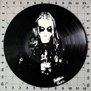 May include: Black vinyl record featuring a black and white portrait of a person with long hair and face paint. The image is centered on the record, with the grooves of the vinyl clearly visible. The record is a standard 12 inches in diameter.