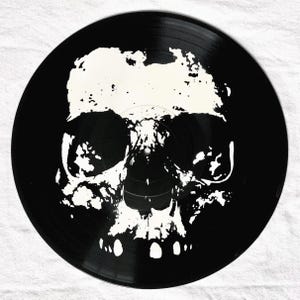 May include: A black vinyl record featuring a white, stylized skull design. The skull's features are created with a textured, almost splattered effect, giving it a unique, artistic appearance. The record is circular and set against a white background.