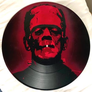May include: A black vinyl record featuring a red and black image of Frankenstein's monster. The artwork depicts the monster's face in a stylized, graphic design. The record has a glossy finish and is a unique piece of art.