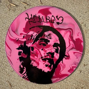 May include: A circular vinyl record featuring a pink and magenta marbled background. A black stencil-style portrait of a person with facial tattoos is centered on the disc. The text HELLBOY is printed in a stylized black font at the top of the record.