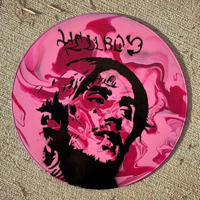 May include: A circular vinyl record featuring a pink and magenta marbled background. A black stencil-style portrait of a person with facial tattoos is centered on the disc. The text HELLBOY is printed in a stylized black font at the top of the record.