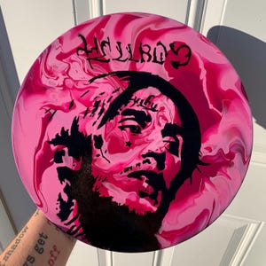 May include: A circular disc with a swirling pink and magenta marbled background. A black silhouette portrait of a person with facial tattoos is centered on the disc. The text HELLBOY is printed in a stylized black font at the top of the disc.