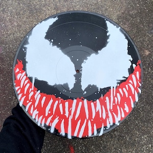 May include: A black vinyl record featuring a white and red painted design. The design depicts a stylized face with white eyes and a red, jagged mouth. The record has a glossy finish and is set against a gray background.