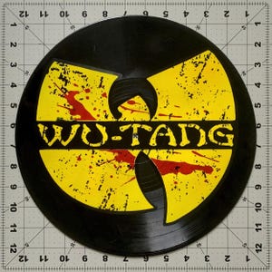 May include: A black vinyl record featuring the Wu-Tang Clan logo in yellow with red splatter details. The text "WU-TANG" is prominently displayed in a bold, yellow font. The record has visible grooves and a circular shape.