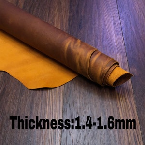May include: A roll of brown leather with a yellow-orange hue. The leather is rolled up and has a rough texture. The text "Thickness: 1.4-1.6mm" is printed on a wooden surface below the leather.
