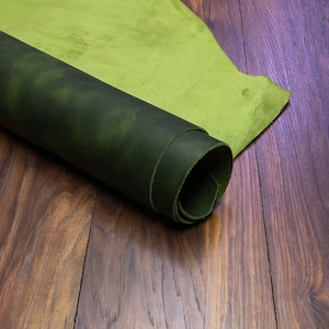 May include: A rolled up piece of dark green leather with a lighter green leather piece partially rolled up on top of it. Both pieces of leather are on a wooden surface.