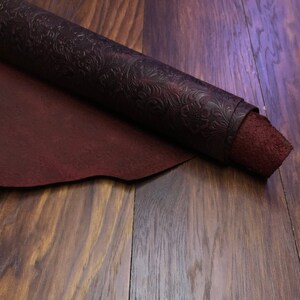 May include: A roll of dark red leather with an embossed floral pattern. The leather is rolled up and lying on a wooden surface.