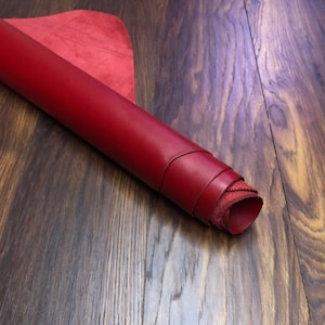 May include: Rolled-up red leather sheet with a smooth, glossy finish. The edges reveal the rough, unfinished underside of the leather. A separate piece of red leather is visible in the background. The leather is displayed on a wooden surface.