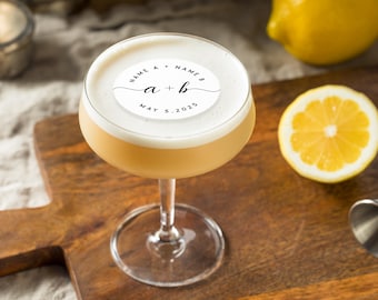 May include: A cocktail in a stemmed glass with a white foam top and a circular label that reads "NAME A + NAME B" and "a + b" with the date "MAY 5, 2025". A lemon half is on a wooden cutting board.