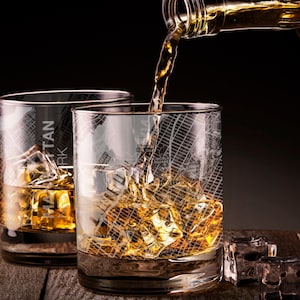May include: Two clear whiskey glasses with a map design, filled with ice and amber liquid. A bottle pours more liquid into one glass. The glasses sit on a wooden surface against a dark background.