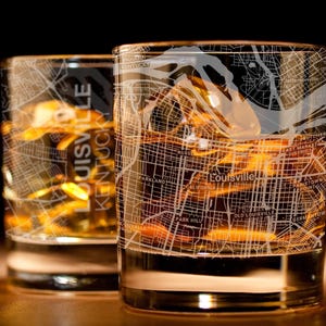 May include: Two clear whiskey glasses filled with ice and amber liquid. The glasses are etched with a detailed map of Louisville, Kentucky, with street names visible. The text "Louisville Kentucky" is vertically printed on one glass.