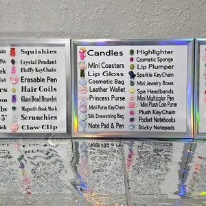 May include: Three silver-framed display boards featuring lists of various small items like acrylic keychains, squishies, candles, and hair clips, each accompanied by a small decorative charm. The boards are arranged side-by-side on a reflective surface.