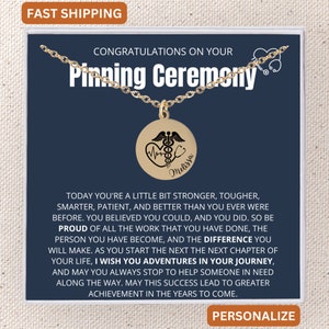 May include: A gold necklace with a round pendant featuring a caduceus medical symbol and a heartbeat. The pendant is engraved with the name "Melissa". The necklace is displayed in a blue box with the text "Congratulations on your Pinning Ceremony".