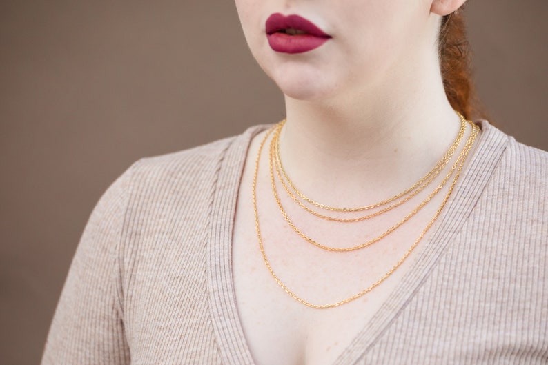 May include: A close-up of a person wearing a beige sweater with four gold chain necklaces layered on top of each other.