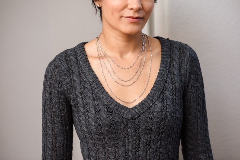 May include: A woman wearing a gray cable knit sweater with a layered silver chain necklace.