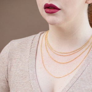 May include: A woman wearing a beige sweater with four gold chain necklaces layered on top of each other.