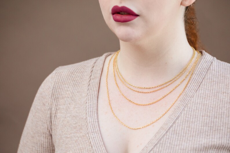 May include: A woman wearing a beige sweater with four gold chain necklaces layered on top of each other.