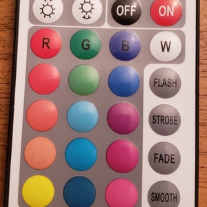 May include: A white remote control with black buttons. The buttons are labeled "ON", "OFF", "R", "G", "B", "W", "FLASH", "STROBE", "FADE", and "SMOOTH". There are also 12 round colored buttons.