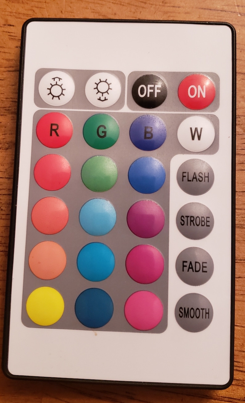 May include: A white remote control with black buttons. The buttons are labeled "ON", "OFF", "R", "G", "B", "W", "FLASH", "STROBE", "FADE", and "SMOOTH". There are also 12 round colored buttons.