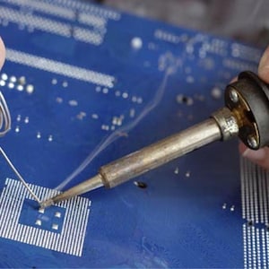 May include: Close-up of a person soldering a circuit board. A soldering iron is held in one hand, while the other hand holds a coil of solder wire. The circuit board is blue with white components.