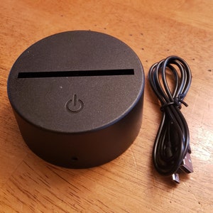 May include: A black circular LED light base with a USB cable. The base has a power button and a slot for inserting a design.