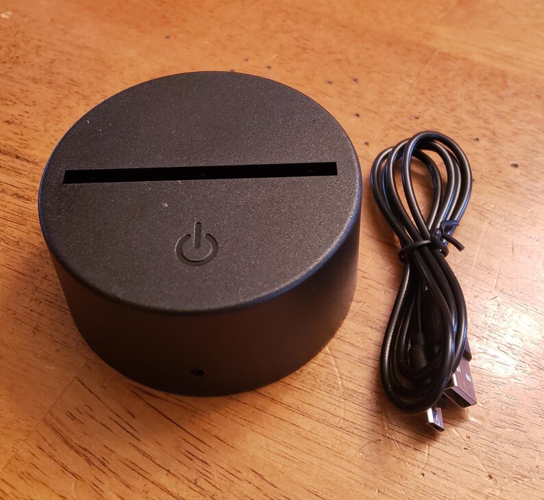 May include: A black circular LED light base with a USB cable. The base has a power button and a slot for inserting a design.