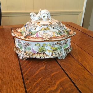 May include: A white porcelain trinket box with a lid featuring a gold-trimmed design of cherubs and floral motifs. The box has a decorative handle on the lid.
