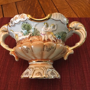 May include: A white ceramic vase with gold accents and a scene of two cherubs in a garden. The vase has two handles and a round base.