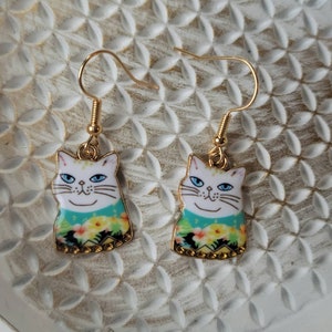 May include: A pair of gold cat earrings with white faces and blue eyes. The cats are wearing a floral patterned shirt with green, yellow, and black accents.