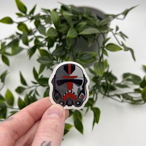 May include: A white, red, and gray Star Wars clone trooper helmet sticker. The helmet has a red stripe down the middle and a black visor.