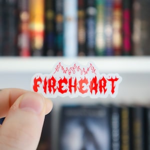 以下が含まれることがあります： 炎が文字の上に描かれた赤い「FIREHEART」という文字が書かれた白いステッカー。ステッカーは本棚の前にある手で持たれている。