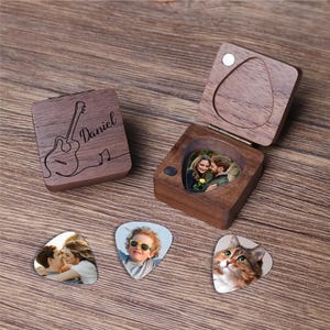 Può includere: Un cofanetto in legno per plettri per chitarra con un contorno di chitarra e il nome "Daniel" inciso sul coperchio. La scatola è aperta, rivelando una foto di una coppia. Sono visibili anche tre plettri per chitarra con foto di una coppia, un bambino e un gatto.