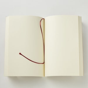 Pu&ograve; includere: Un taccuino aperto con pagine bianche color avorio e un sottile segnalibro a nastro rosso scuro che fuoriesce dal dorso centrale. Il taccuino &egrave; appoggiato in piano, mostrando due pagine vuote con superfici in carta opaca liscia e bordi netti.