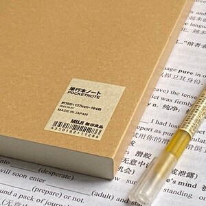 Pu&ograve; includere: Un taccuino color marrone chiaro con un'etichetta rettangolare riportante il testo POCKETNOTE, 195x137mm, 184 fogli, MADE IN JAPAN e MUJI. Una penna dorata con cappuccio trasparente poggia accanto al taccuino su un foglio con testo stampato.