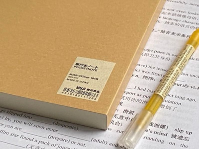 Pu&ograve; includere: Un taccuino color marrone chiaro con un'etichetta rettangolare riportante il testo POCKETNOTE, 195x137mm, 184 fogli, MADE IN JAPAN e MUJI. Una penna dorata con cappuccio trasparente poggia accanto al taccuino su un foglio con testo stampato.