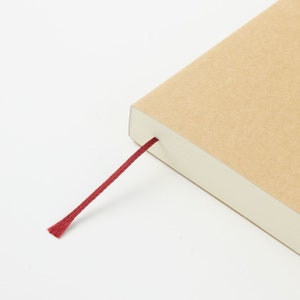 Pu&ograve; includere: Un taccuino con una copertina in cartone color marrone chiaro e un sottile segnalibro a nastro rosso scuro che fuoriesce dal dorso. Le pagine sono bianche e impilate in modo ordinato, mostrando un bordo netto su sfondo bianco.
