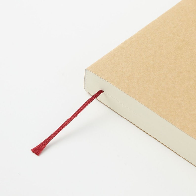 Pu&ograve; includere: Un taccuino con una copertina in cartone color marrone chiaro e un sottile segnalibro a nastro rosso scuro che fuoriesce dal dorso. Le pagine sono bianche e impilate in modo ordinato, mostrando un bordo netto su sfondo bianco.