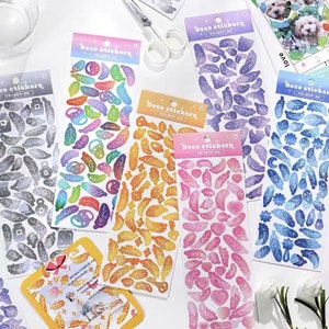 Könnte beinhalten: Mehrere Bögen mit dekorativen Aufklebern mit Feder- und Blattmotiven in verschiedenen Farben wie Lila, Orange, Rosa und Blau. Jedes Blatt trägt die Aufschrift "Deco Stickers" und enthält eine Vielzahl von Formen und Größen für Bastelarbeiten und Scrapbooking.