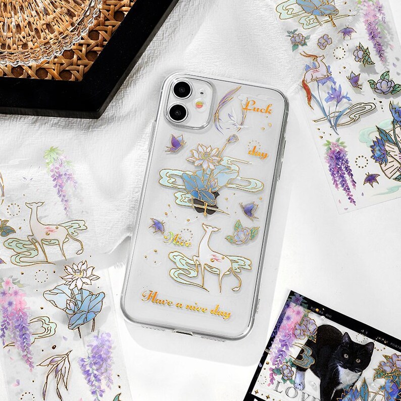 Pu&ograve; includere: Una custodia per telefono trasparente con un design colorato che presenta cervi, fiori e nuvole. La custodia ha il testo "Luck day" e "Have a nice day" stampato su di essa.