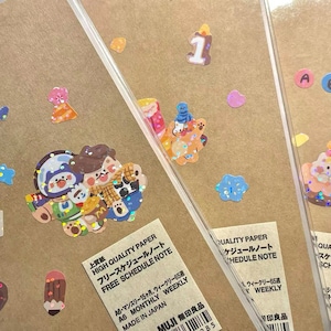 May include: Three brown paper notebooks with colorful stickers on the front. The stickers include a birthday cake, a number one, and a group of cartoon animals. The notebooks are labeled "High Quality Paper Free Schedule Note" and "A6 Monthly Weekly Made in Japan".