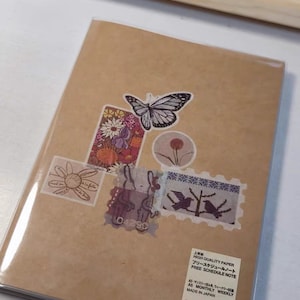 May include: A brown notebook with a clear plastic cover. The cover has several colorful stickers on it, including a butterfly, flowers, and a bunny. The notebook is labeled "High Quality Paper Free Schedule Note" and "AS Monthly Weekly" in Japanese. The notebook is made in Japan and is a MUJI product.