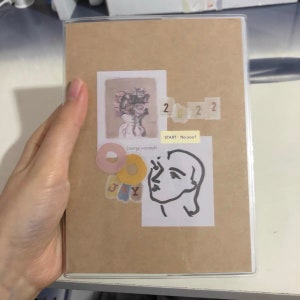 May include: A brown notebook with a clear plastic cover. The notebook has various stickers on it, including a floral illustration, a black and white line drawing of a face, and a sticker with the text "START: No.0001".