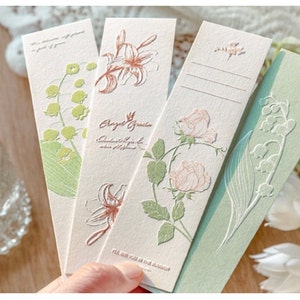 Könnte beinhalten: Vier Blumen-Lesezeichen mit geprägten Designs. Die Lesezeichen zeigen eine Lilie, eine Rose und zwei verschiedene Designs von Maiglöckchen. Der Text auf einem Lesezeichen lautet "Angel Garden, Cultivate your life with happiness".