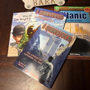 May include: A stack of children's books featuring the "I Survived" series, including titles about the September 11 attacks, the Titanic, and the Grizzly bears. The books are on a dark wooden surface.