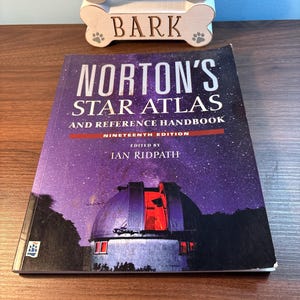 May include: A copy of "Norton's Star Atlas and Reference Handbook" with a purple and black cover featuring a night sky and an observatory. The book is the nineteenth edition, edited by Ian Ridpath. A wooden dog bone-shaped sign that says "Love Bark" is in the background.