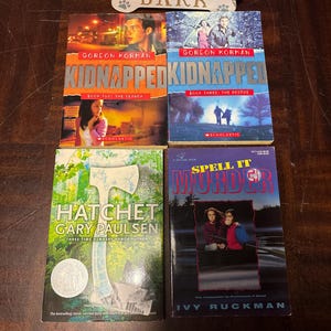 May include: Four paperback books are displayed. Two books are titled "Kidnapped" by Gordon Korman, one "Hatchet" by Gary Paulsen, and one "Spell It Murder" by Ivy Ruckman. The books are on a dark wooden surface, with a decorative wooden sign that says "Love Bark".