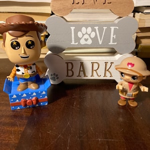 May include: Two figurines of cartoon characters, one resembling Woody from Toy Story, and another in safari attire, are displayed. Behind them are three wooden bone-shaped signs that read "LIVE", "LOVE" with a paw print, and "BARK".